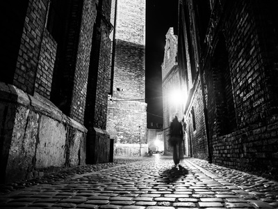 Illuminated cobbled street with light reflections on cobblestones in old historical city by night. Dark blurred silhouette of person evokes Jack the Ripper. Black and white image.